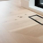Aria vent floor register with black frame installed flush in light oak hardwood floor, modern Toronto living room with grey sectional sofa and large windows in background